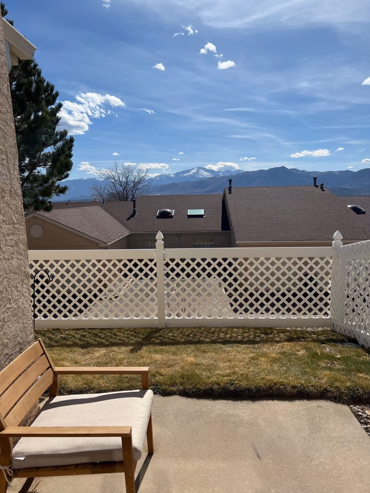 Outdoor patio area with a wooden chair featuring a beige cushion, a small patch of grass, and a white lattice fence. In the background, there are rooftops of neighboring buildings and a scenic view of mountains under a blue sky with scattered clouds.