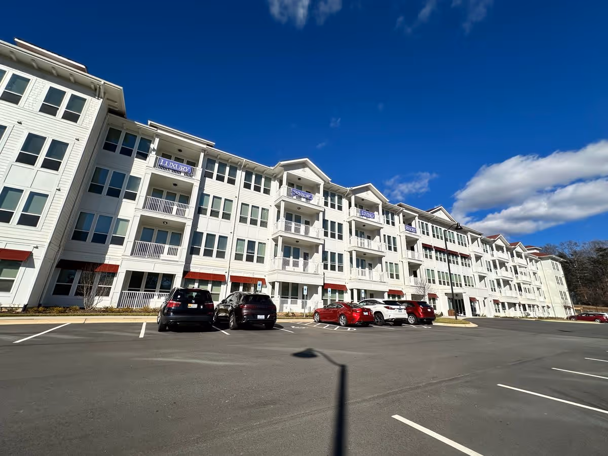 Wide-angle view of a white multi-story senior living building with balconies and a parking lot under a blue sky.