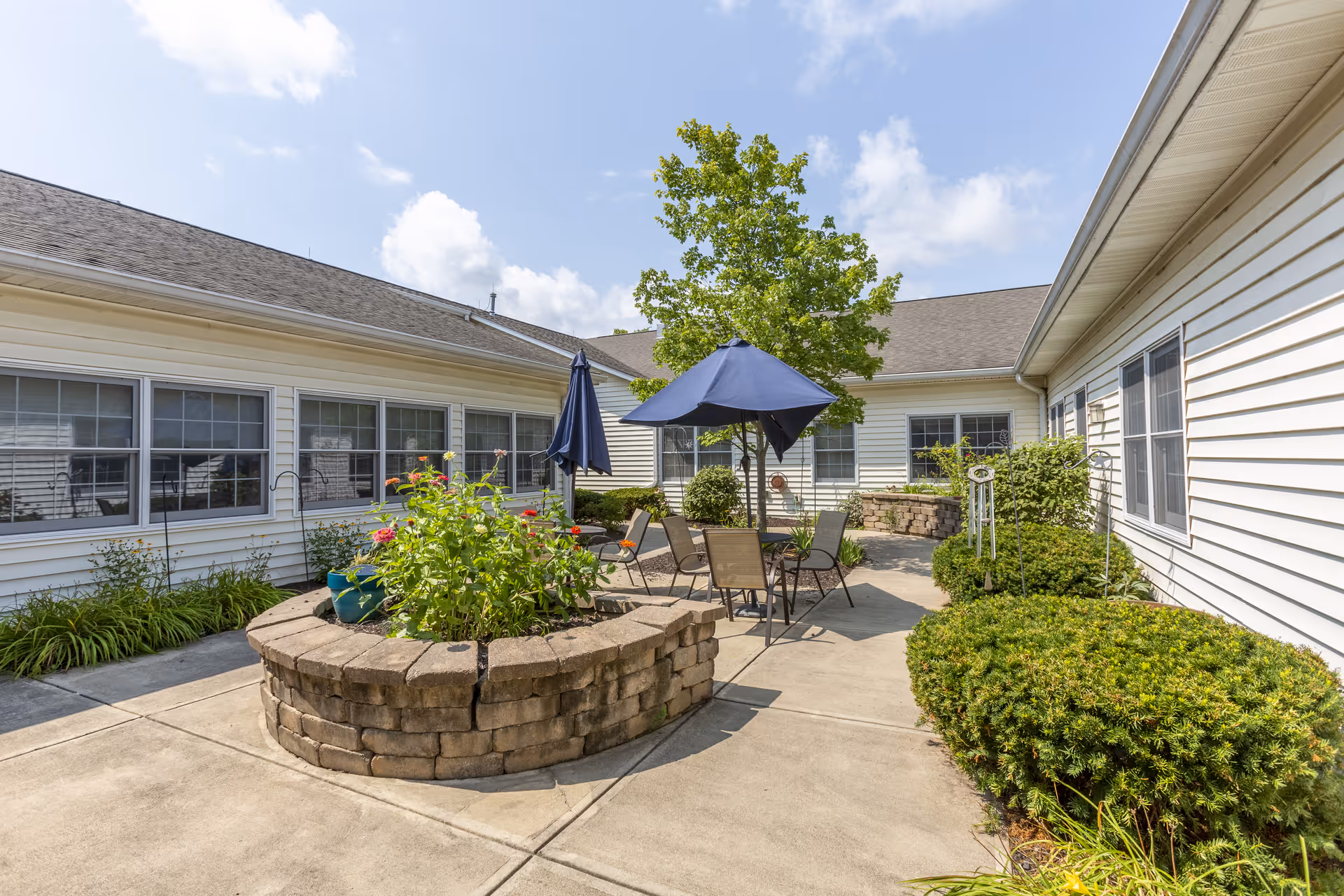 Outdoor courtyard area at Grand Victorian of Greenwood with a circular raised brick planter filled with flowers, patio tables and chairs with blue umbrellas, surrounded by white buildings and green bushes under a partly cloudy sky.