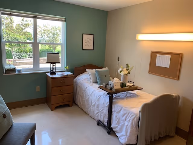 A small, neatly arranged bedroom with a single bed covered in a white quilt and several pillows. Next to the bed is a wooden nightstand with a lamp and a small plant. A window with a view of greenery and a bench outside lets in natural light. A framed quote hangs on the green accent wall, and a corkboard is mounted on the adjacent beige wall. A rolling overbed table with a tissue box, flowers, and other items is positioned over the bed. A cushioned chair is partially visible in the foreground.