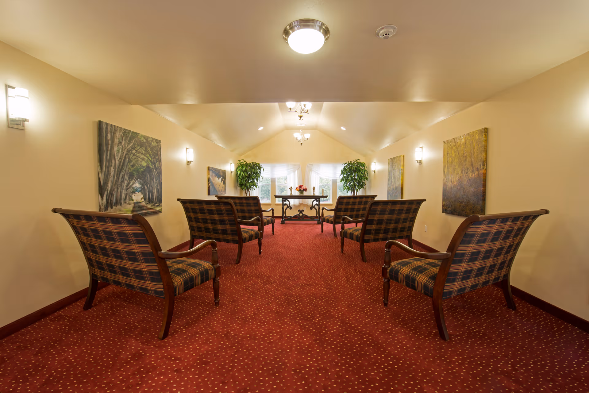 A cozy sitting area with six wooden armchairs upholstered in a plaid fabric arranged in two rows facing a small table with flowers. The room has a red carpet with a subtle pattern, cream-colored walls adorned with framed landscape paintings, and two large potted plants near a window with sheer white curtains. The ceiling is vaulted with multiple light fixtures providing warm lighting.