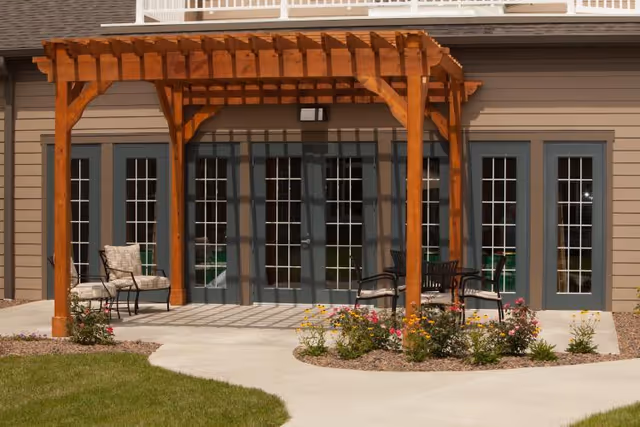 Outdoor patio area with a wooden pergola attached to a building featuring multiple glass-paneled doors. There are chairs arranged under the pergola and flower beds with blooming flowers along the concrete walkway.