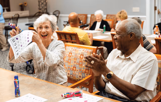 Two elderly people sitting at a table playing bingo, one woman holding up her bingo card and smiling excitedly while a man claps his hands. Other seniors are seated at tables in the background in a communal room.