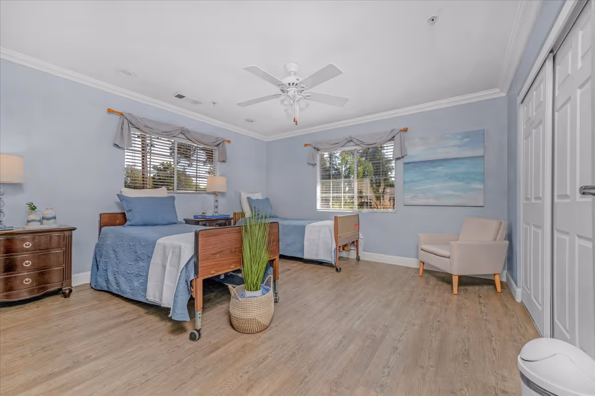 A bright and spacious bedroom with two single beds featuring blue bedding and wooden headboards. The room has light blue walls, two windows with blinds and valances, a ceiling fan, a wooden nightstand with a lamp and decorative items, a beige armchair, a large closet with white sliding doors, and a potted plant on the floor.