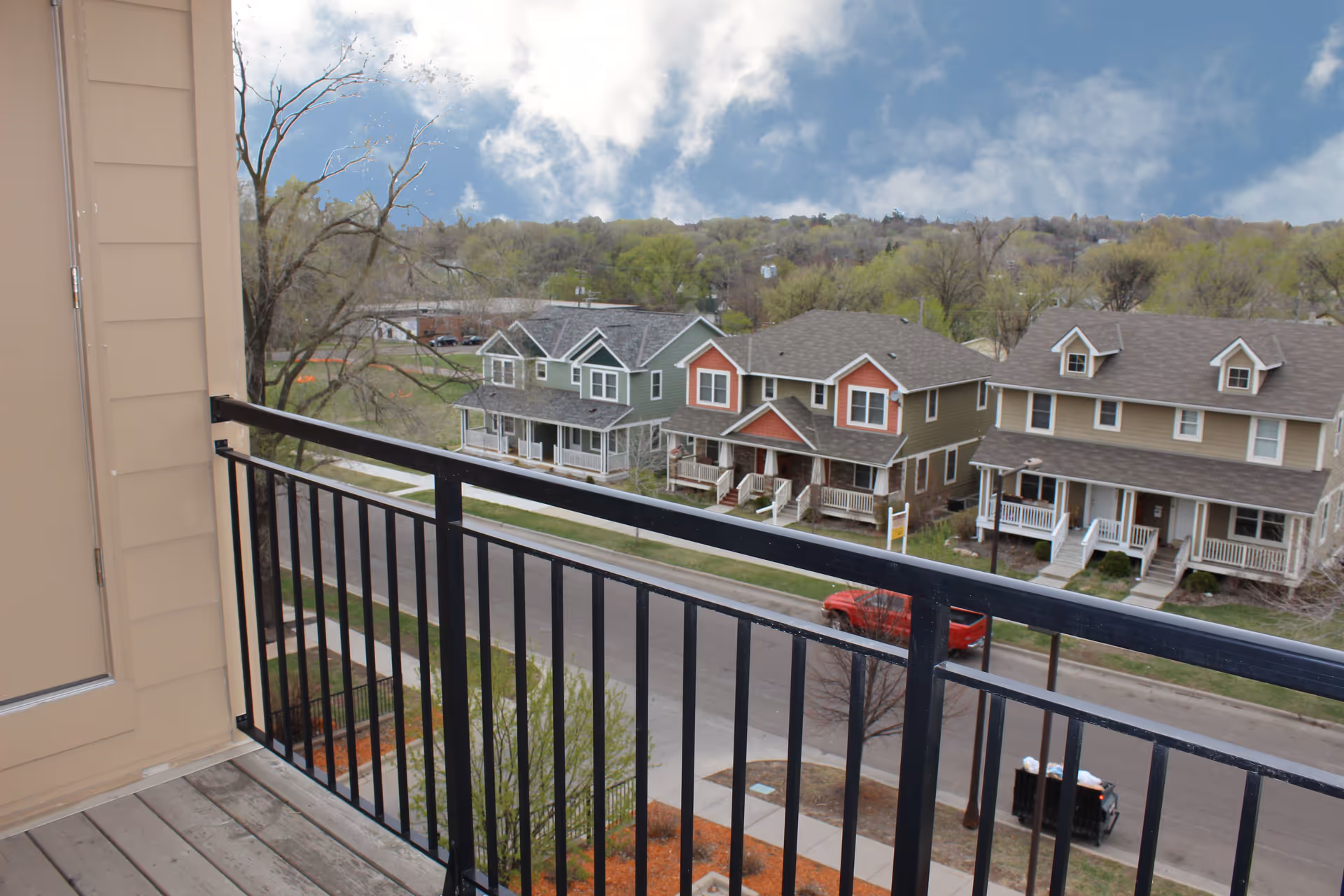 View from a balcony with black metal railing overlooking a street with several two-story houses and trees in the background under a partly cloudy sky.