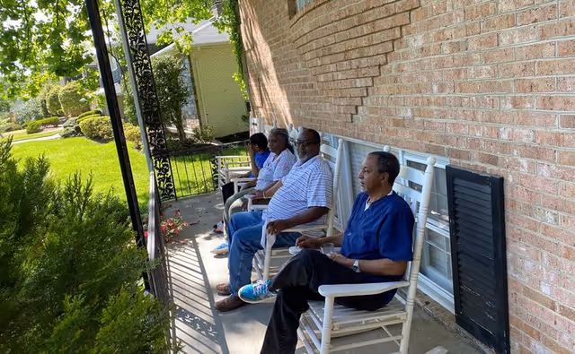 Four elderly individuals sitting on white rocking chairs on a shaded porch next to a brick building, with greenery and a lawn visible in the background.