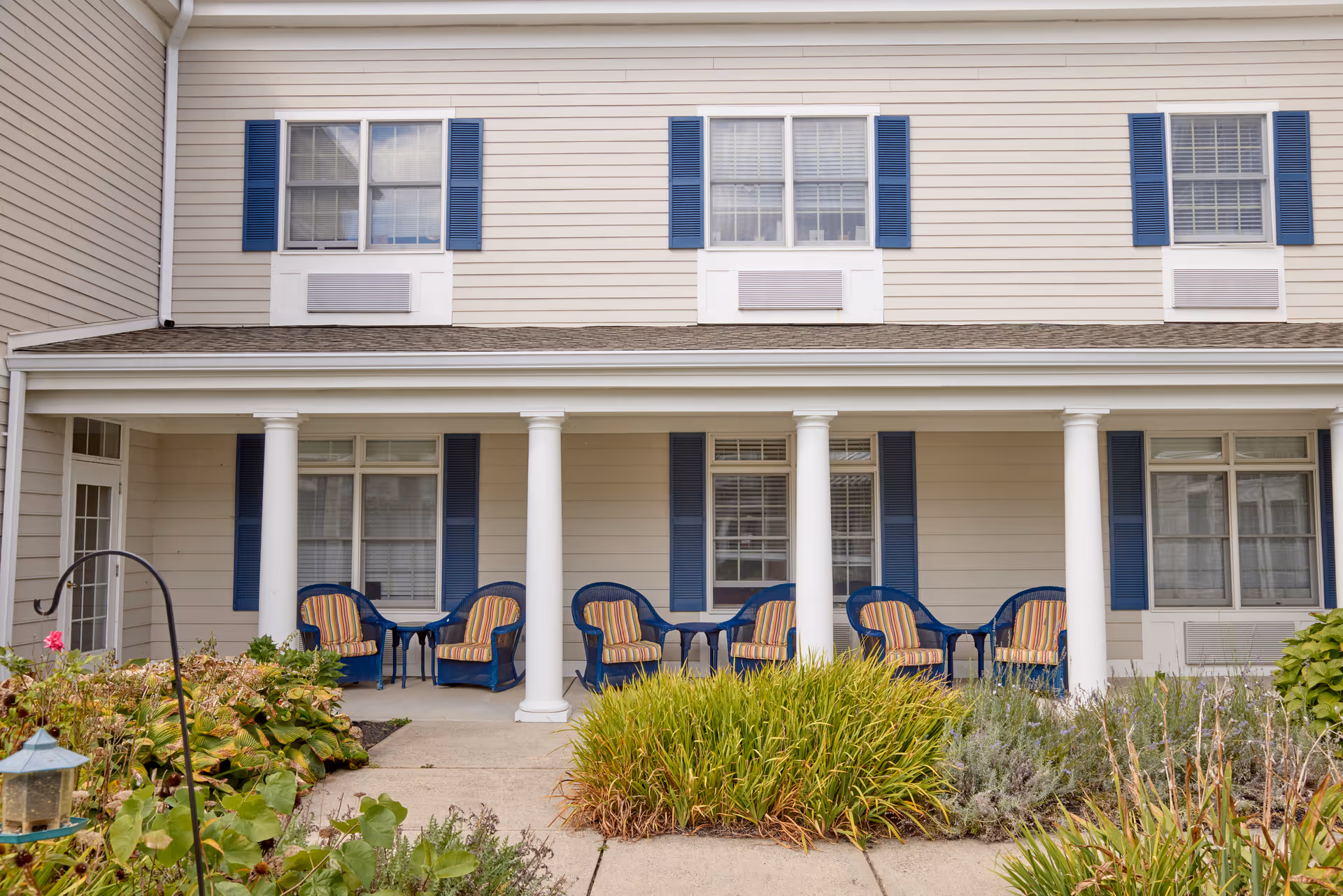 Front porch of a senior living building with white columns, blue chairs with striped cushions, blue shutters and landscaped walkway.