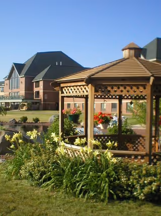 Wooden gazebo surrounded by flowering plants with the senior living building in the background under a clear blue sky.