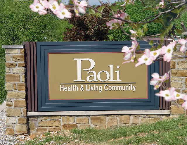 Stone-framed entrance sign reading "Paoli Health & Living Community" with flowering branches in the foreground.