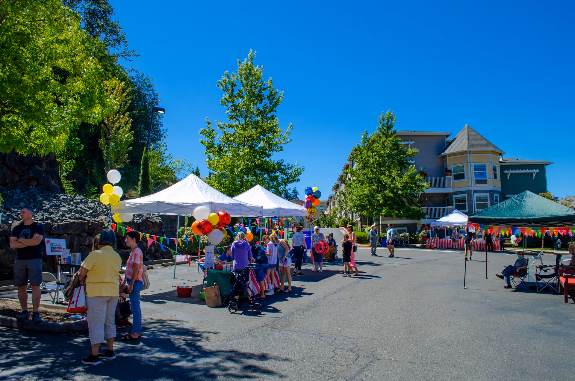 Outdoor community event at Chateau Bothell Landing Retirement with people gathered around white tents decorated with colorful balloons and pennant banners on a sunny day. Trees and a multi-story building are visible in the background.