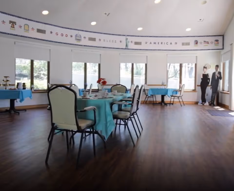 A bright dining room with several tables covered in blue tablecloths and set with cups and plates. The room has large windows with white blinds, wooden flooring, and a patriotic banner on the wall that reads 'God Bless America.' There are two life-sized cutouts of a man and woman dressed formally near the windows.
