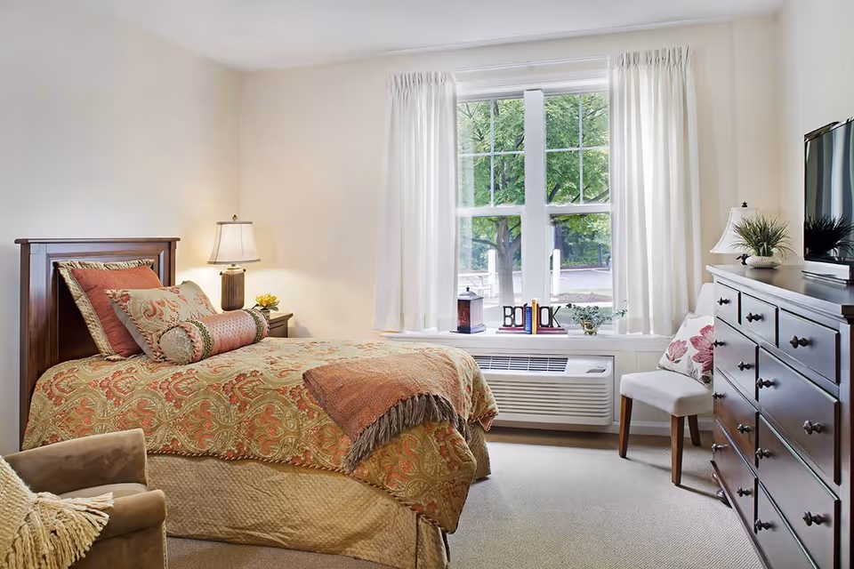 A cozy bedroom with a single bed covered in a patterned bedspread and multiple pillows. There is a wooden nightstand with a lamp and a small flower arrangement next to the bed. A large window with white curtains lets in natural light, and a white chair with a floral pillow sits beside a dark wooden dresser with a flat-screen TV on top. A beige armchair with a throw blanket is partially visible in the foreground.