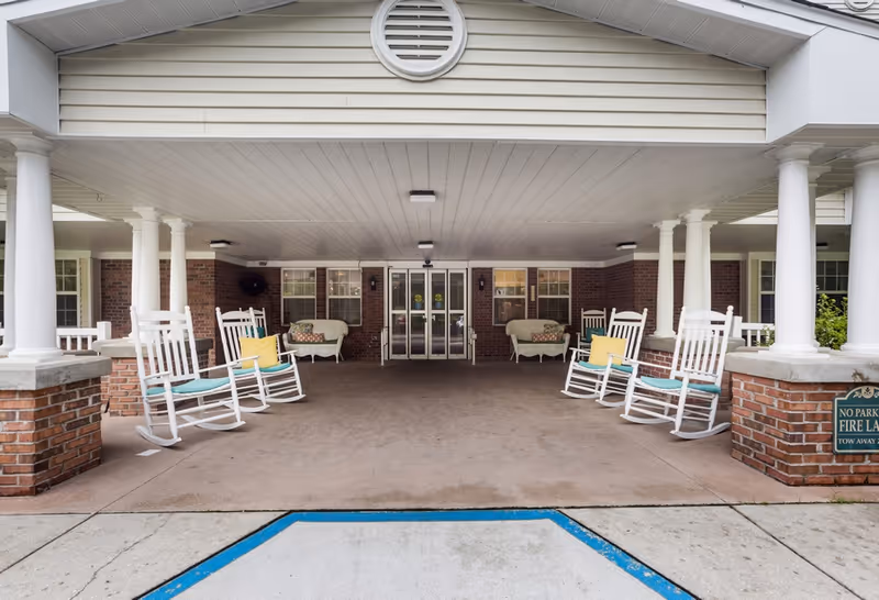Covered entrance area of a senior living facility with white rocking chairs and cushioned benches arranged on either side. The building features brick walls, white columns, and double glass doors at the center. A sign indicating no parking in the fire lane is visible on the right side.