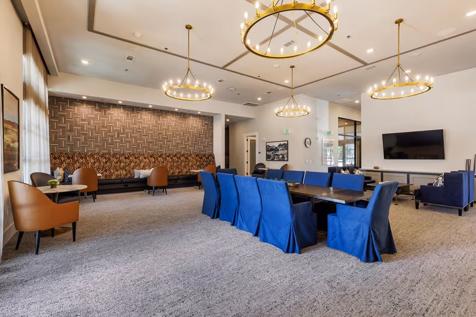 Spacious senior living common room with a long table surrounded by blue chairs, additional lounge seating, chandeliers, and a wall-mounted TV.