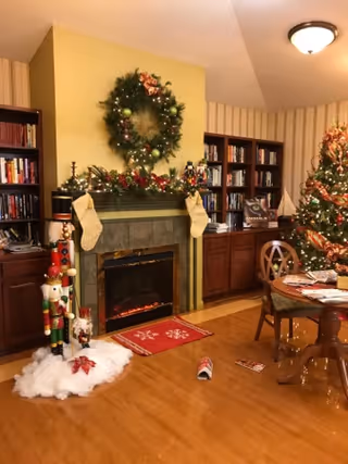 A cozy living room decorated for Christmas with a lit fireplace, a large wreath above the mantel, stockings hanging on the mantel, a decorated Christmas tree, a nutcracker figure, bookshelves filled with books, and a round wooden table with chairs.