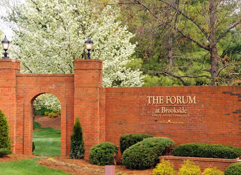 Brick entrance wall with an arched doorway and two lamp posts on top, surrounded by green bushes and trees with white blossoms. The wall has a sign that reads 'THE FORUM at Brookside Senior Living Community'.