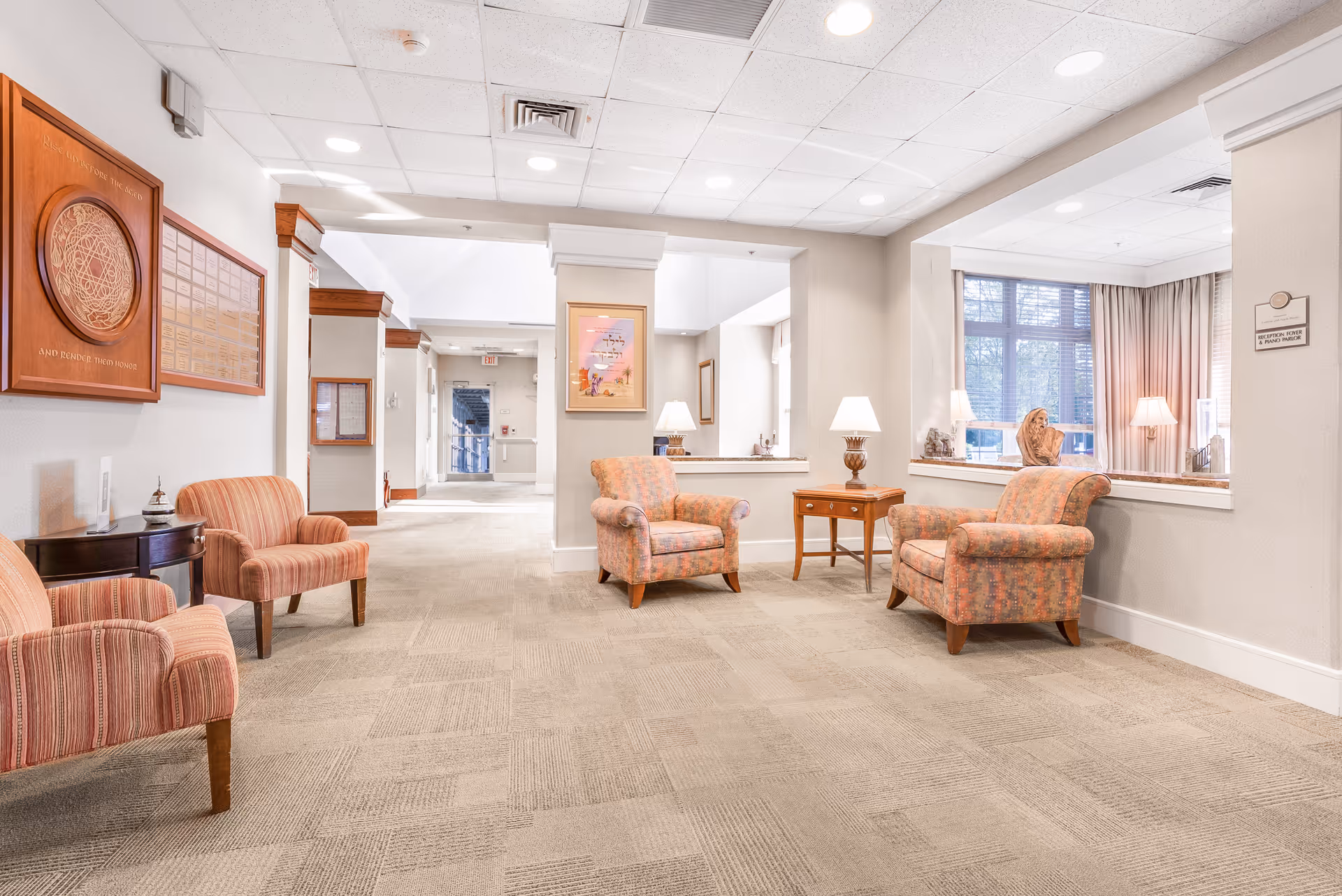 Well-lit senior living facility seating area with upholstered armchairs, side tables and lamps in a carpeted lobby.