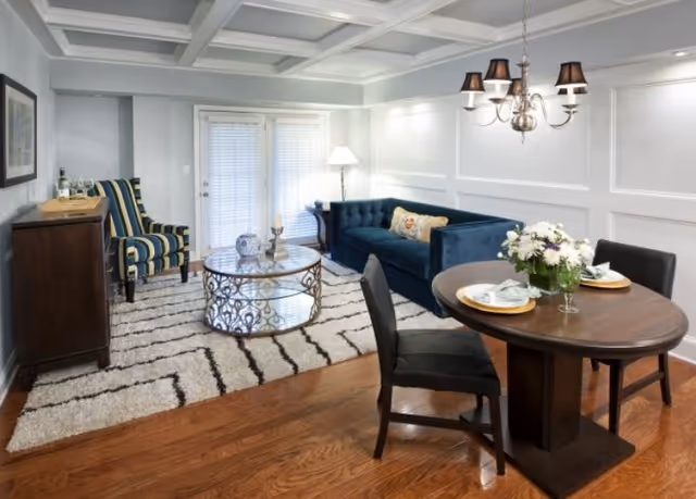 A cozy living and dining area with a blue velvet sofa, a striped armchair, a round glass coffee table on a patterned rug, and a small wooden dining table set for two with a flower centerpiece. The room has white paneled walls, a coffered ceiling, and wooden flooring.