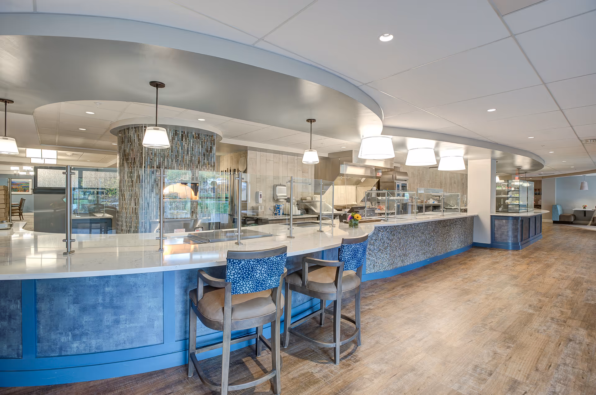 A modern dining area with a long counter featuring protective glass partitions and several high chairs with blue patterned backs. The space has a curved ceiling design with hanging pendant lights and a tiled column in the background. The floor is wooden, and the area appears clean and well-lit.