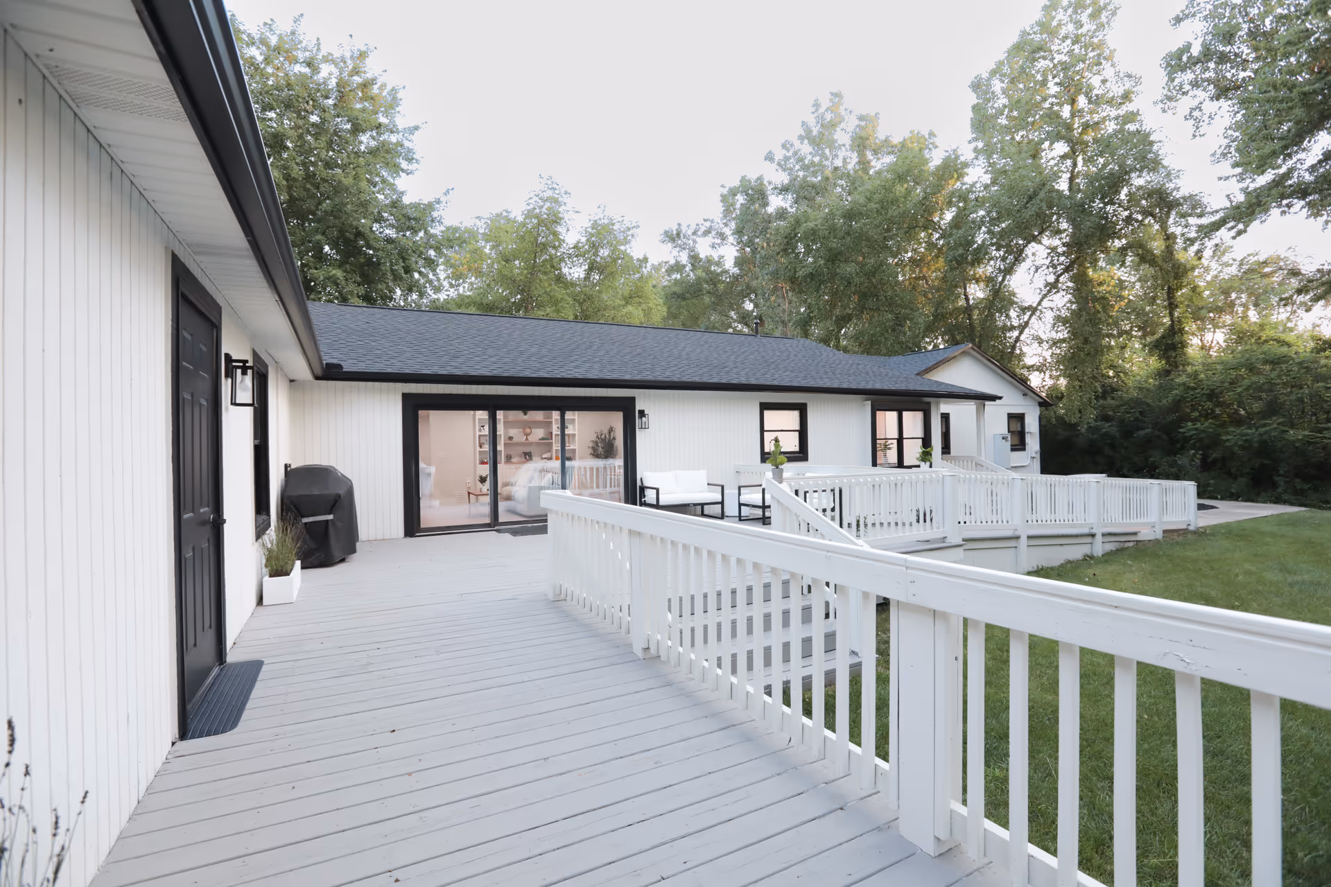 Outdoor view of a senior living facility showing a wide wooden deck with white railings and a ramp leading to the entrance. The building exterior is white with black trim around the doors and windows. There are some outdoor chairs and a grill on the deck, with green grass and trees surrounding the area.