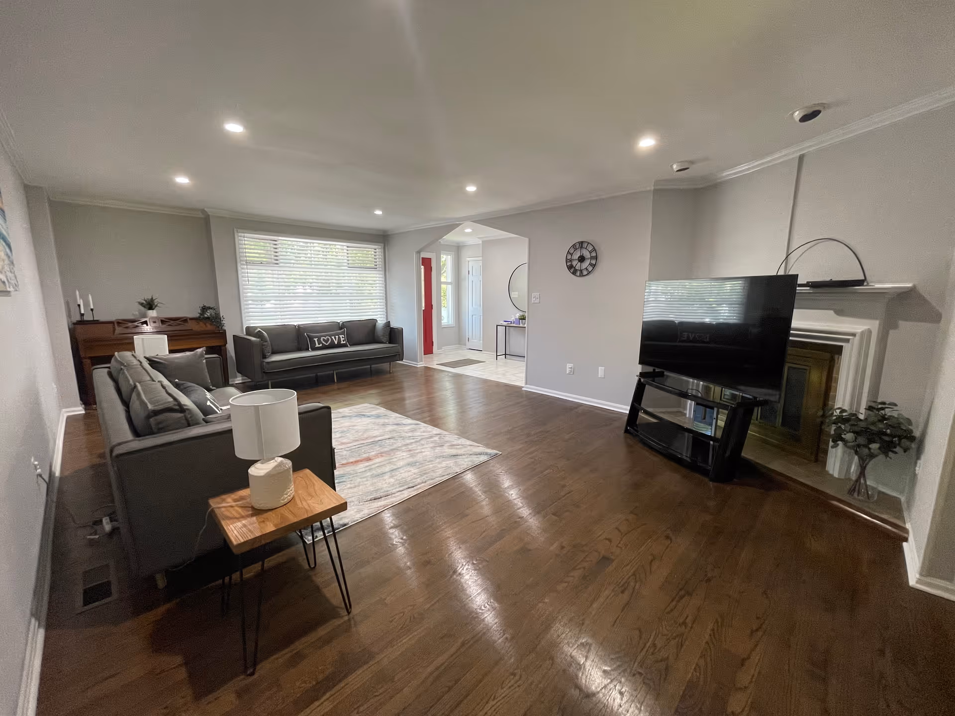 A spacious living room with wooden floors, two gray sofas, a small wooden side table with a lamp, a piano against the wall, a large window with blinds, a flat-screen TV on a stand in front of a fireplace, and a wall clock. The room is well-lit with recessed ceiling lights.
