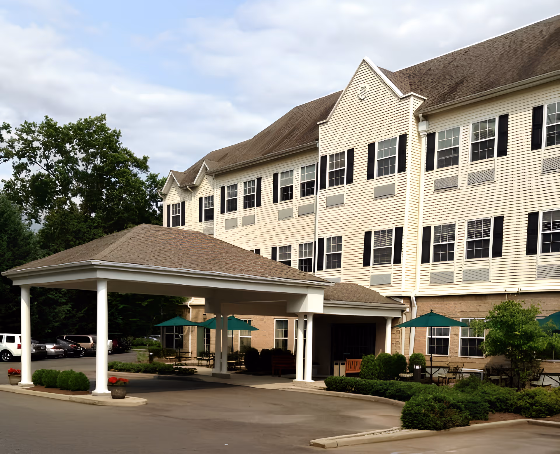 Exterior view of Avalon Assisted Living at Bridgewater, showing a large multi-story building with many windows and black shutters. The entrance has a covered drop-off area supported by white columns. There are green umbrellas and outdoor seating visible near the entrance, with some landscaping and parked cars in the background.