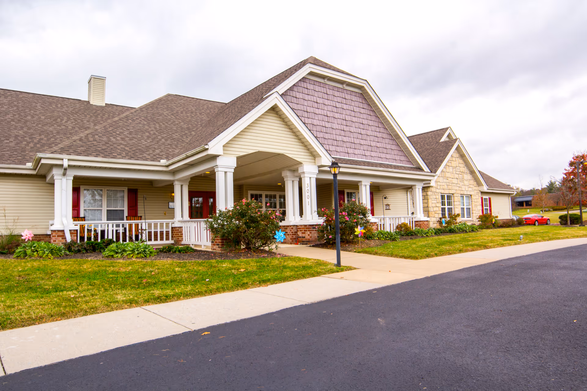 Exterior view of a single-story senior living facility building with beige siding, a brown shingled roof, white columns, and a covered entrance. There are bushes and flowers planted along the front, a sidewalk leading to the entrance, and a red car parked in the distance on the right. The sky is overcast.