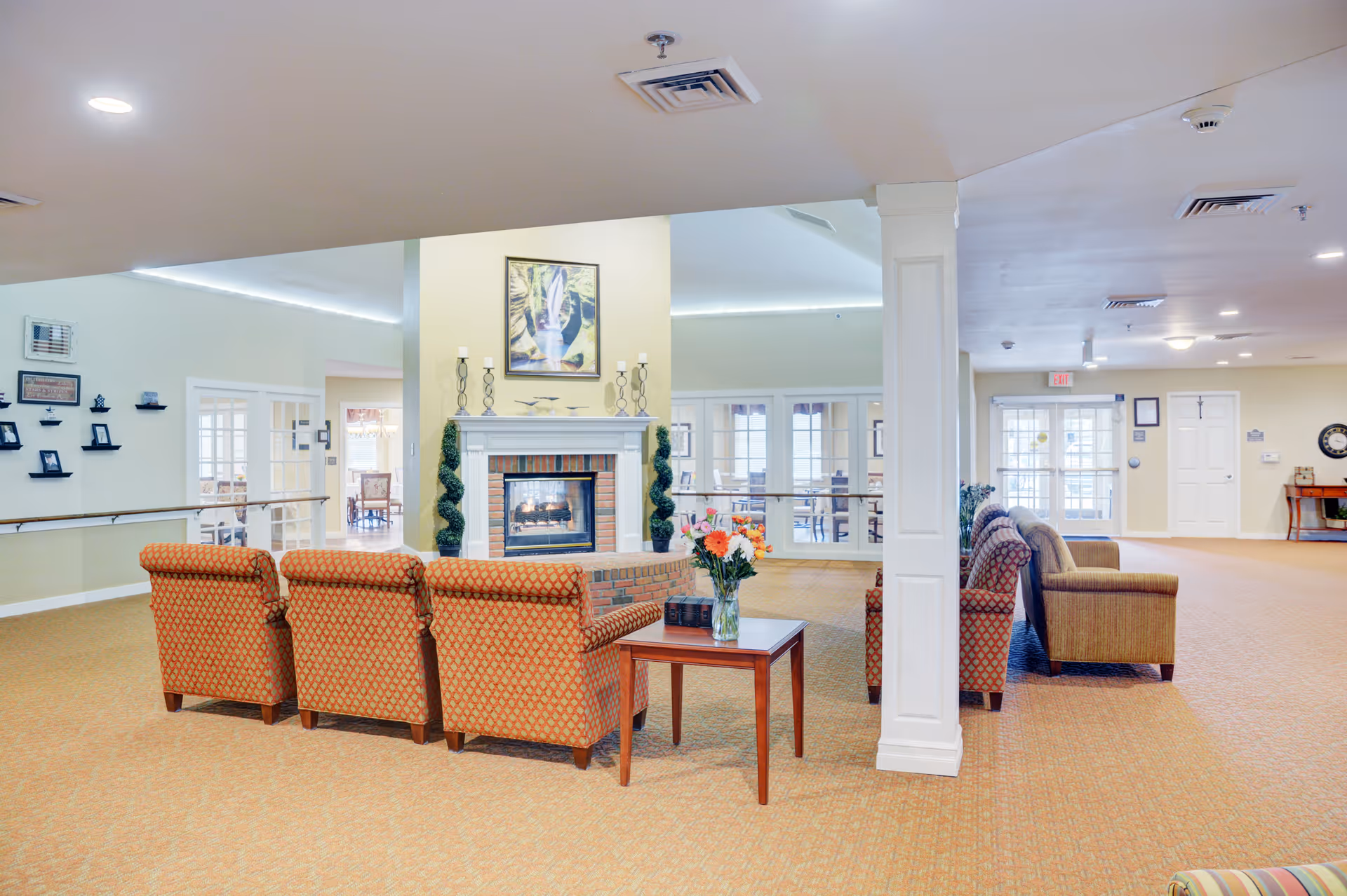 A spacious and well-lit common area in a senior living facility featuring a central fireplace with decorative candles and plants on the mantel. Surrounding the fireplace are several upholstered armchairs arranged for socializing. A small wooden table with a vase of flowers is placed near the chairs. The room has beige walls, carpeted floors, and large windows and doors in the background allowing natural light to enter. The ceiling has recessed lighting and ventilation fixtures.