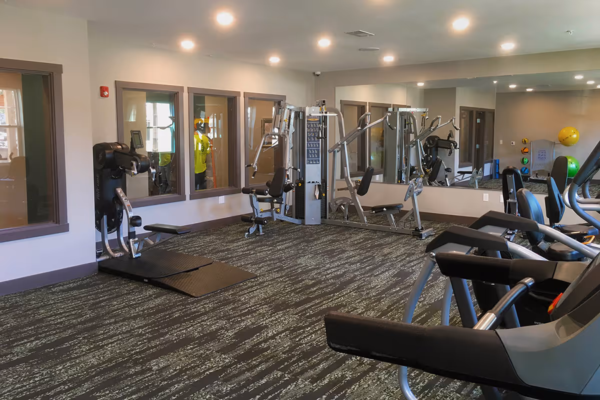 Interior view of a fitness room with various exercise equipment including weight machines, treadmills, and exercise balls. The room has large windows, mirrors on one wall, and recessed lighting in the ceiling.