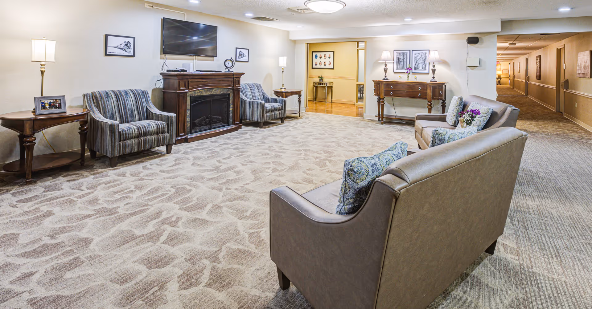 A spacious senior living community lounge with patterned carpet, two striped armchairs, a wooden electric fireplace with a mounted flat-screen TV above it, and two beige sofas with decorative pillows. The room is softly lit with floor lamps and table lamps on wooden side tables and a console table. There is a hallway visible to the right and an open doorway leading to another room in the background.