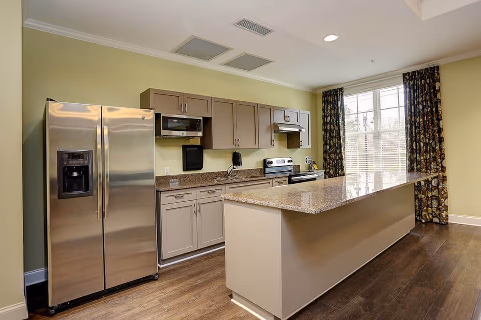 A modern kitchen with stainless steel refrigerator, microwave, stove, and granite countertops. There is a large kitchen island in the center and a window with floral curtains letting in natural light.