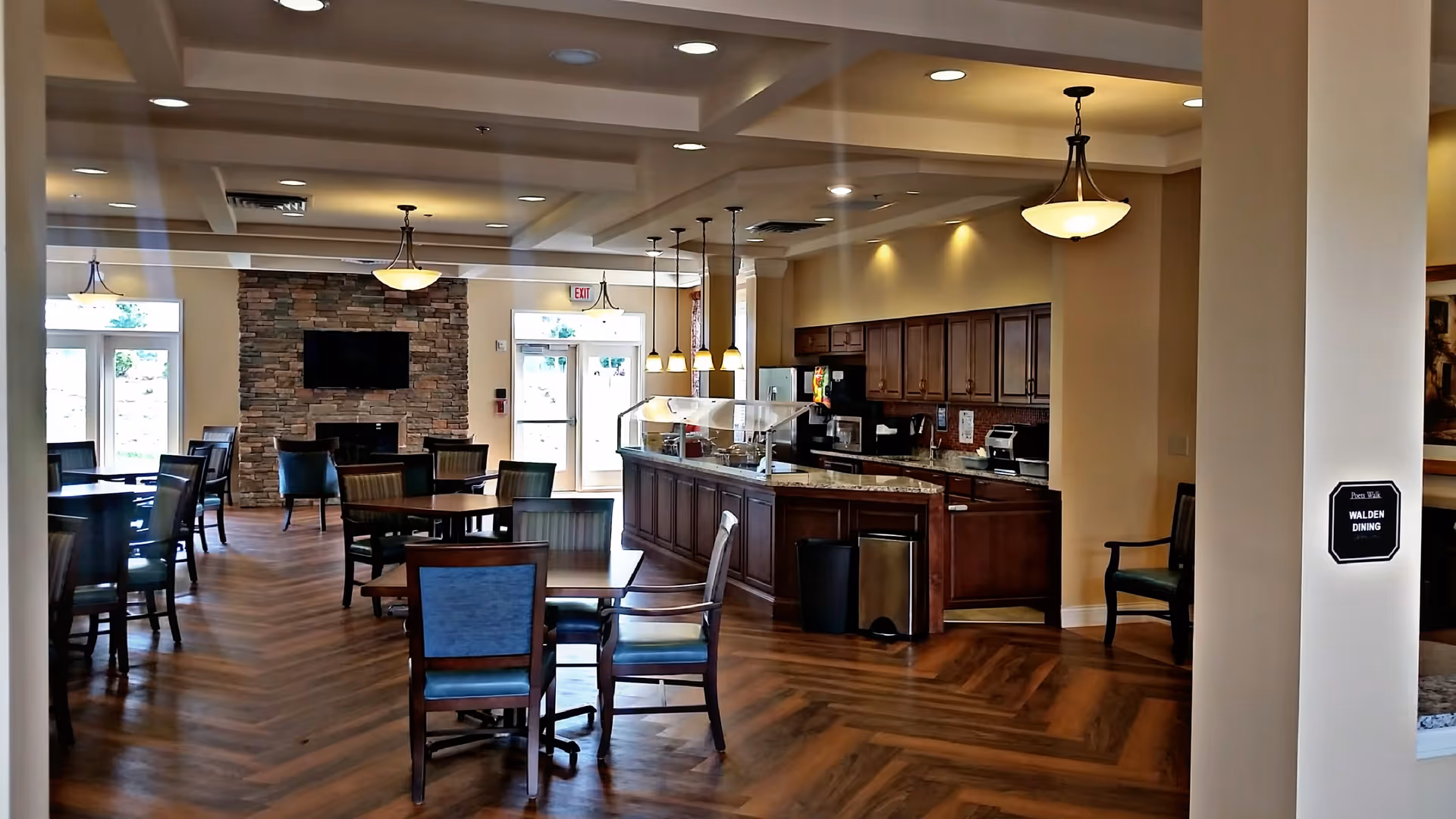 Interior view of a senior living facility dining room with wooden tables and chairs, a serving counter with hanging lights, a stone fireplace with a mounted TV, and large windows letting in natural light.