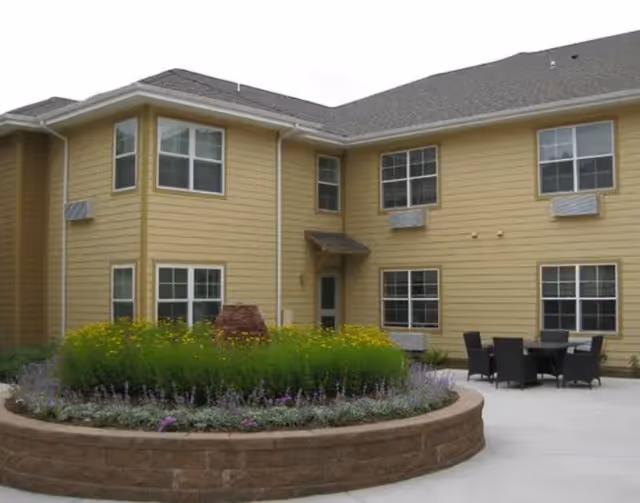 Outdoor courtyard area of a senior living facility with a circular raised flower bed filled with green plants and yellow flowers. The building has beige siding with multiple windows and air conditioning units. There is a patio area with a table and several chairs.