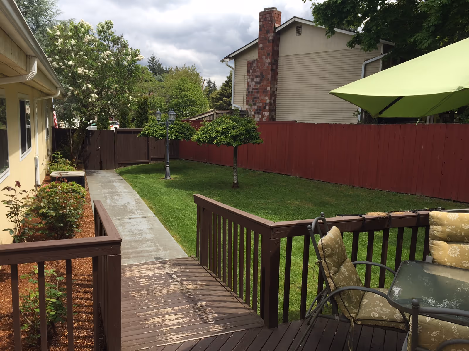 Backyard area of a residential facility with a wooden deck featuring cushioned patio chairs and a glass table under a green umbrella. A concrete pathway leads through a grassy lawn bordered by a red wooden fence and small trees. The side of a yellow building is visible on the left with some plants along the walkway.