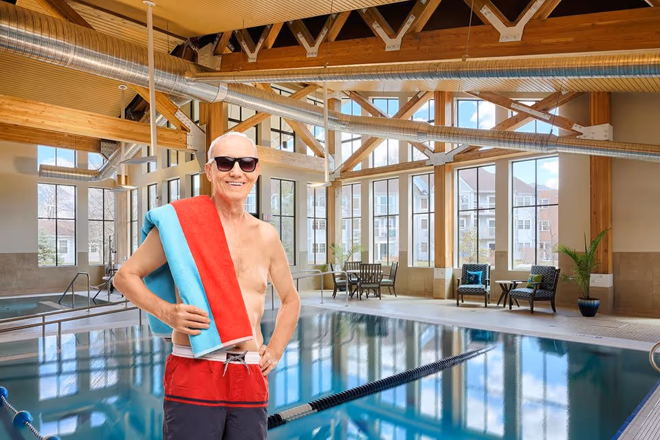 An elderly man wearing sunglasses and red swim trunks stands smiling by an indoor swimming pool, holding a red and blue towel over his shoulder. The pool area features large windows, wooden beams, and modern industrial-style ductwork, with seating and plants visible in the background.
