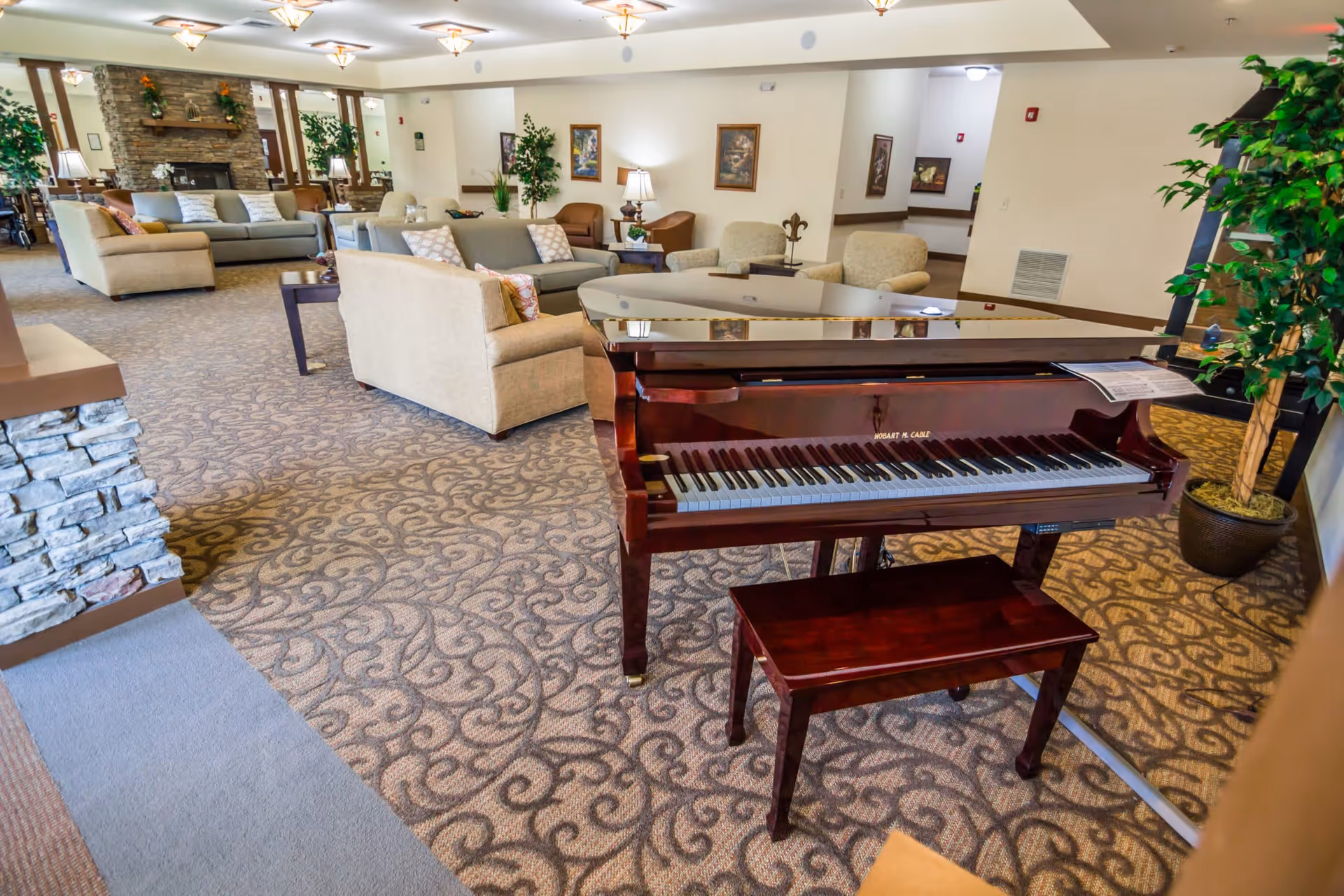 A spacious and well-lit living room area in a senior living facility featuring a polished grand piano with a matching bench in the foreground. The room has patterned carpet flooring, several beige and light-colored sofas and armchairs arranged around coffee tables, potted plants, framed artwork on the walls, and a stone fireplace in the background.