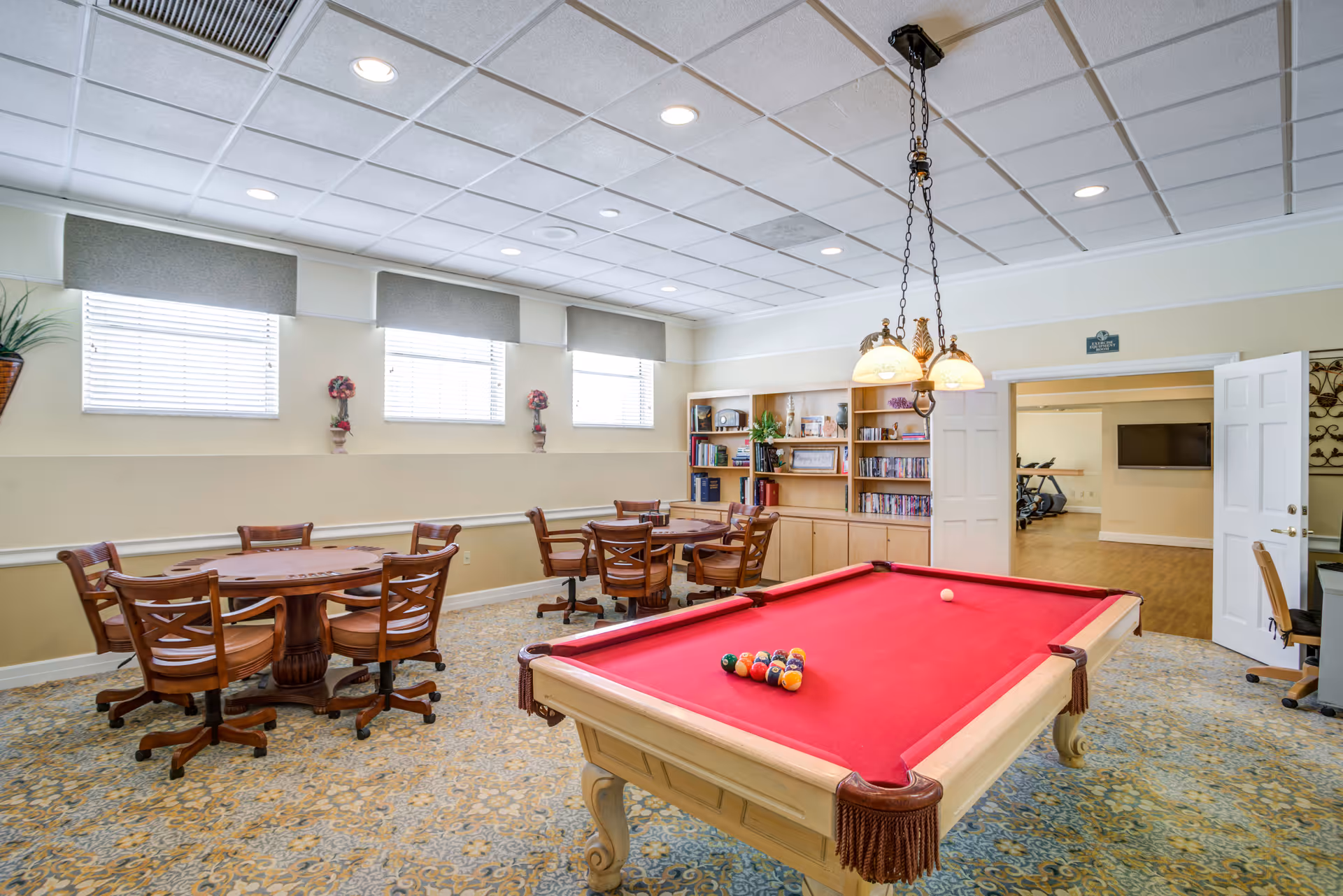 A recreational room with a red felt pool table in the foreground, surrounded by wooden chairs and tables. The room has three windows with blinds and valances, a bookshelf filled with books and decorative items, and a hanging light fixture above the pool table. An open doorway leads to another room with exercise equipment and a wall-mounted TV.