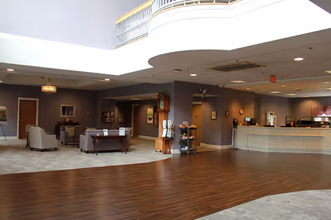 Spacious senior living facility lobby with wood and carpet flooring, seating areas with armchairs and sofas, a reception desk, and soft lighting from ceiling fixtures.