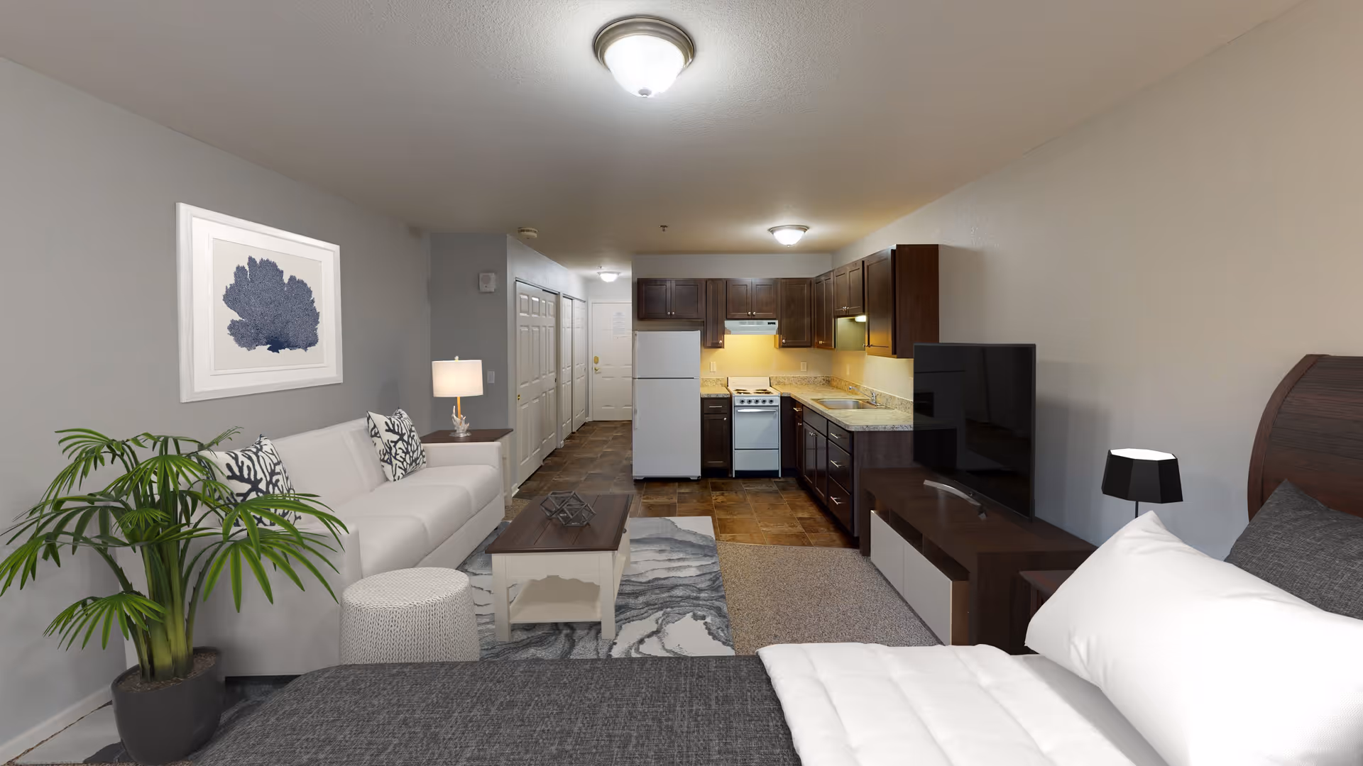 A studio apartment interior featuring a bed with white and gray bedding in the foreground, a dark wood TV stand with a flat-screen TV, a small black lamp, a white sofa with patterned pillows, a coffee table, a potted plant, and a kitchen area with dark wood cabinets, a white refrigerator, and a stove in the background.