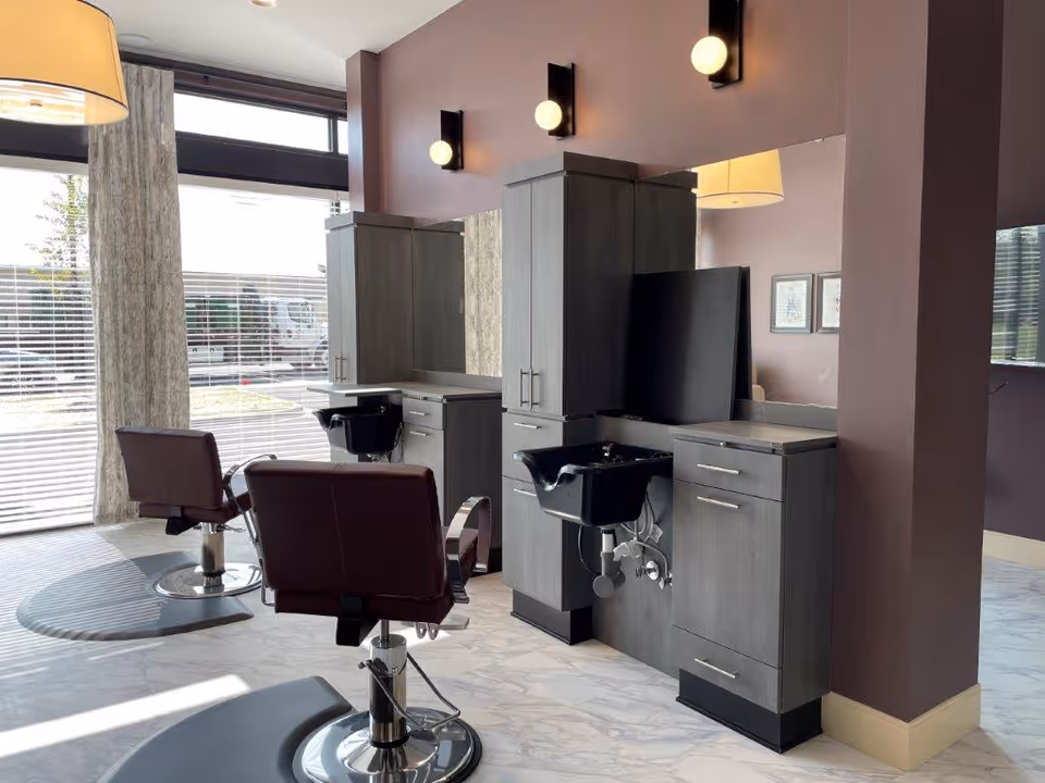 Interior of a salon area with two styling chairs in front of large mirrors and black wash basins attached to gray cabinets. The room has purple walls, large windows with blinds, and modern wall-mounted lights above the mirrors.