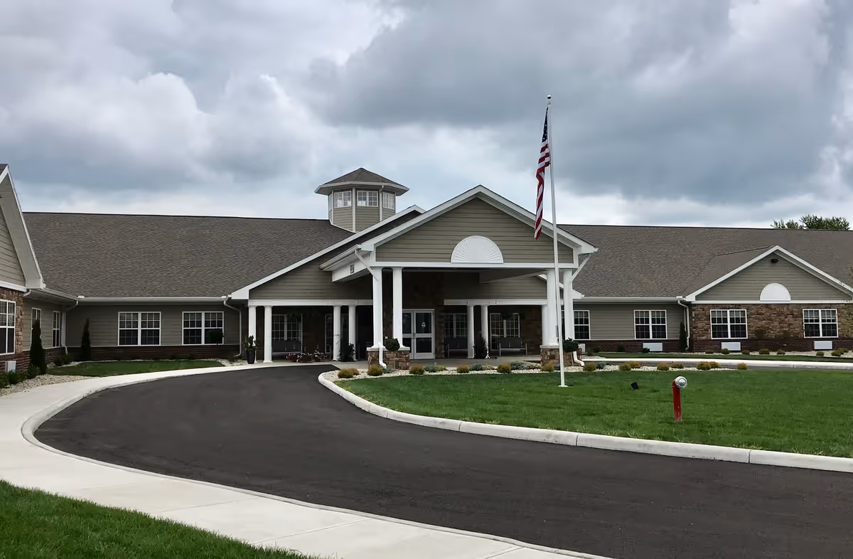 Front exterior view of The Springs of Lima facility showing a single-story building with a covered entrance, an American flag on a flagpole, a circular driveway, and a well-maintained lawn under a cloudy sky.