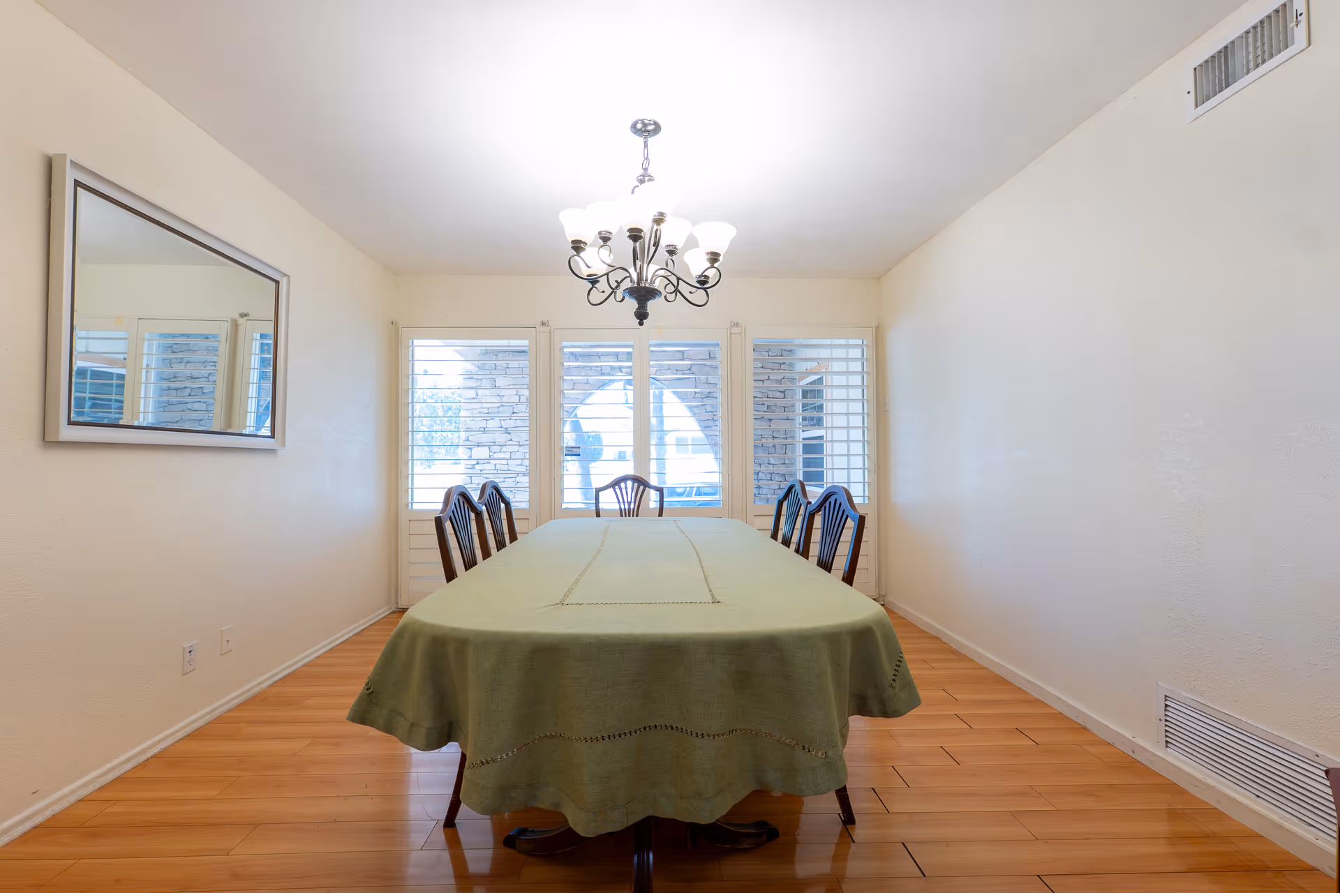 A dining room with a rectangular table covered with a green tablecloth and six wooden chairs. There is a chandelier hanging from the ceiling above the table. Three large windows with white shutters are on the far wall, and a large mirror is mounted on the left wall. The floor is wooden, and the walls are painted white.