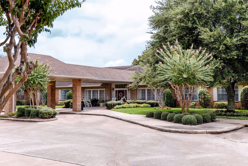Exterior view of a single-story assisted living facility with a covered entrance, brick pillars, and well-maintained landscaping including trimmed bushes and trees under a partly cloudy sky.