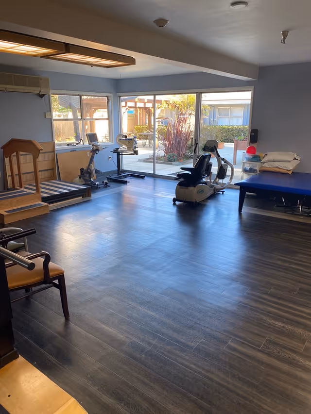 A spacious room with exercise equipment including a stationary bike and a recumbent bike, a padded therapy table with pillows, and a chair with armrests. Large windows and sliding glass doors provide a view of an outdoor patio area with plants and seating. The room has dark wood flooring and light blue walls.