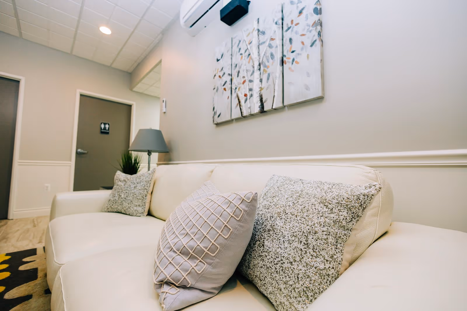 A beige leather couch with three decorative pillows in a softly lit room. Above the couch is a multi-panel wall art featuring abstract tree branches with leaves. To the left, there is a gray door with a restroom sign, a floor lamp, and a small plant on a side table. The walls are painted light beige with white trim, and the ceiling has recessed lighting and a white air conditioning unit.