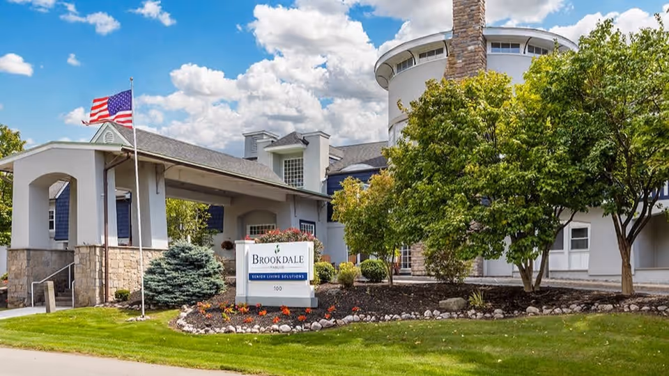 Exterior view of Brookdale Manlius senior living facility showing the entrance with a covered driveway, an American flag on a flagpole, landscaped greenery including trees and shrubs, and a sign that reads 'Brookdale Senior Living Solutions'. The building has a light-colored facade with a stone base and a partly cloudy sky in the background.