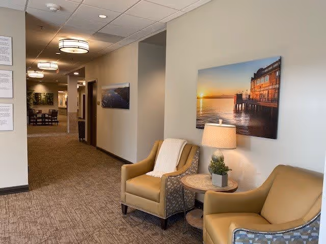 A hallway in a senior living facility with two tan armchairs and a small round table with a lamp and plant between them. The walls are light-colored and decorated with framed photographs of waterfront scenes. The hallway extends into a dining area with tables and chairs visible in the background.