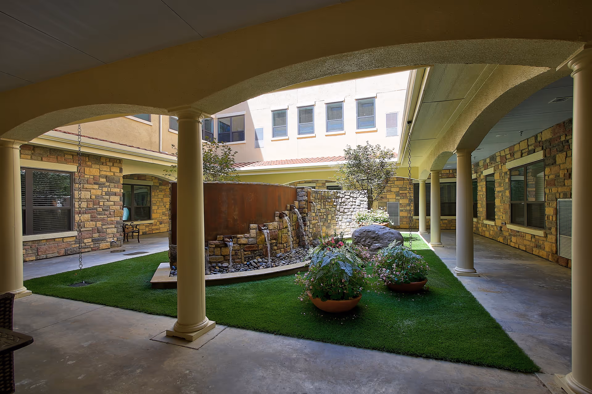 A covered courtyard area with beige columns and arches surrounding a small garden with green grass, potted plants, a large rock, and a stone water feature with multiple spouts. The courtyard is enclosed by a building with stone and stucco walls and several windows.