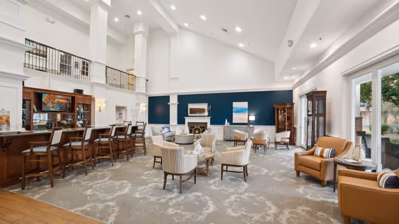 A spacious and well-lit living room area in a senior living facility with high vaulted ceilings, a fireplace, several comfortable chairs arranged around small tables, a wooden bar with bar stools, and large windows letting in natural light.