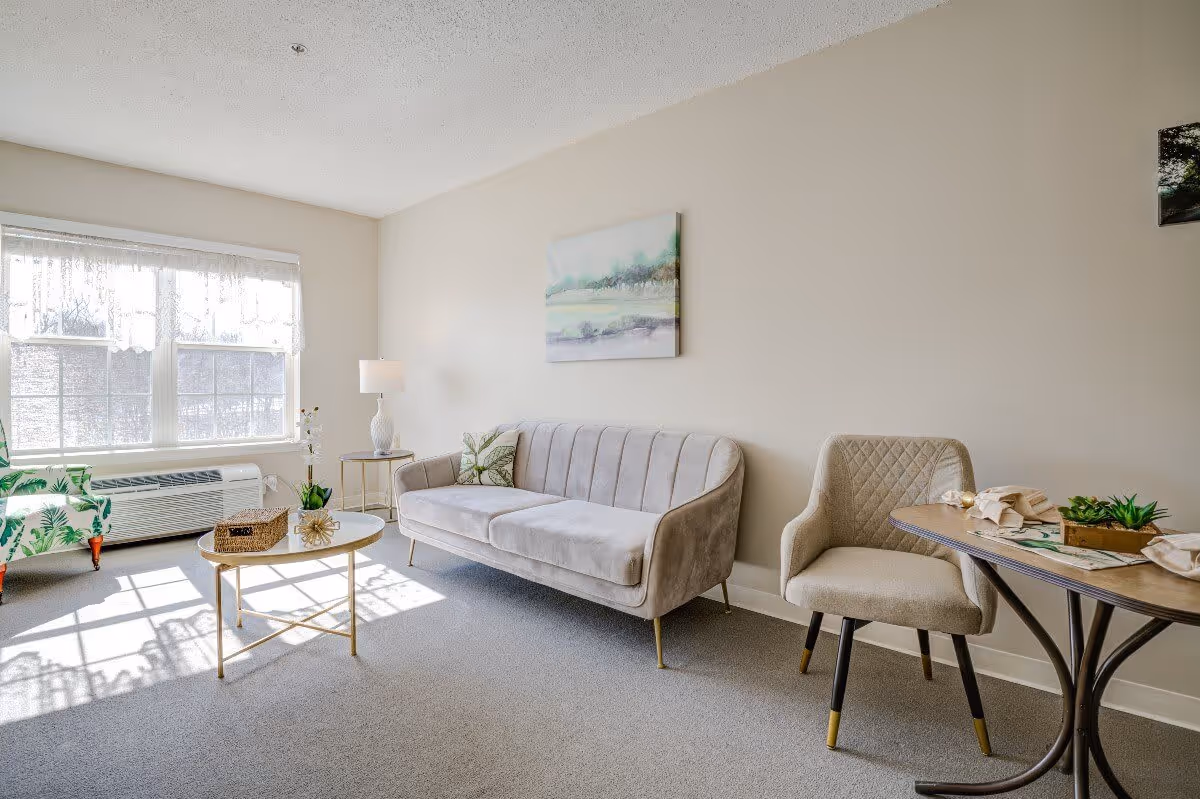 A bright living room with a large window letting in natural light. The room features a beige sofa with a decorative pillow, a beige upholstered chair, a round coffee table with decorative items, a side table with a lamp, and a small dining table set with napkins and a small plant. The walls are light-colored with a landscape painting hanging above the sofa.